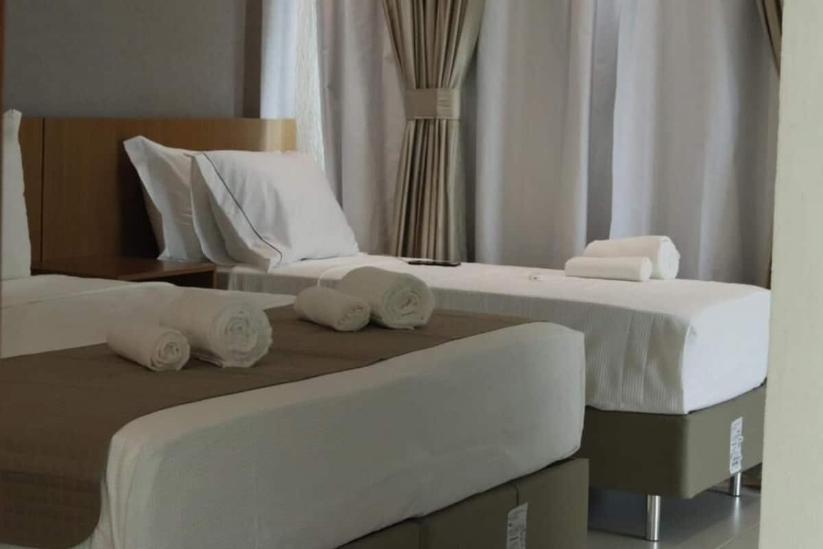 A hotel room with two beds, each with white linens and a brown throw blanket. There are rolled towels on the beds, and the room has a neutral color scheme with curtains.