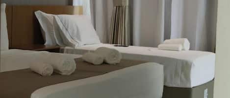 A hotel room with two beds, each with white linens and a brown throw blanket. There are rolled towels on the beds, and the room has a neutral color scheme with curtains.
