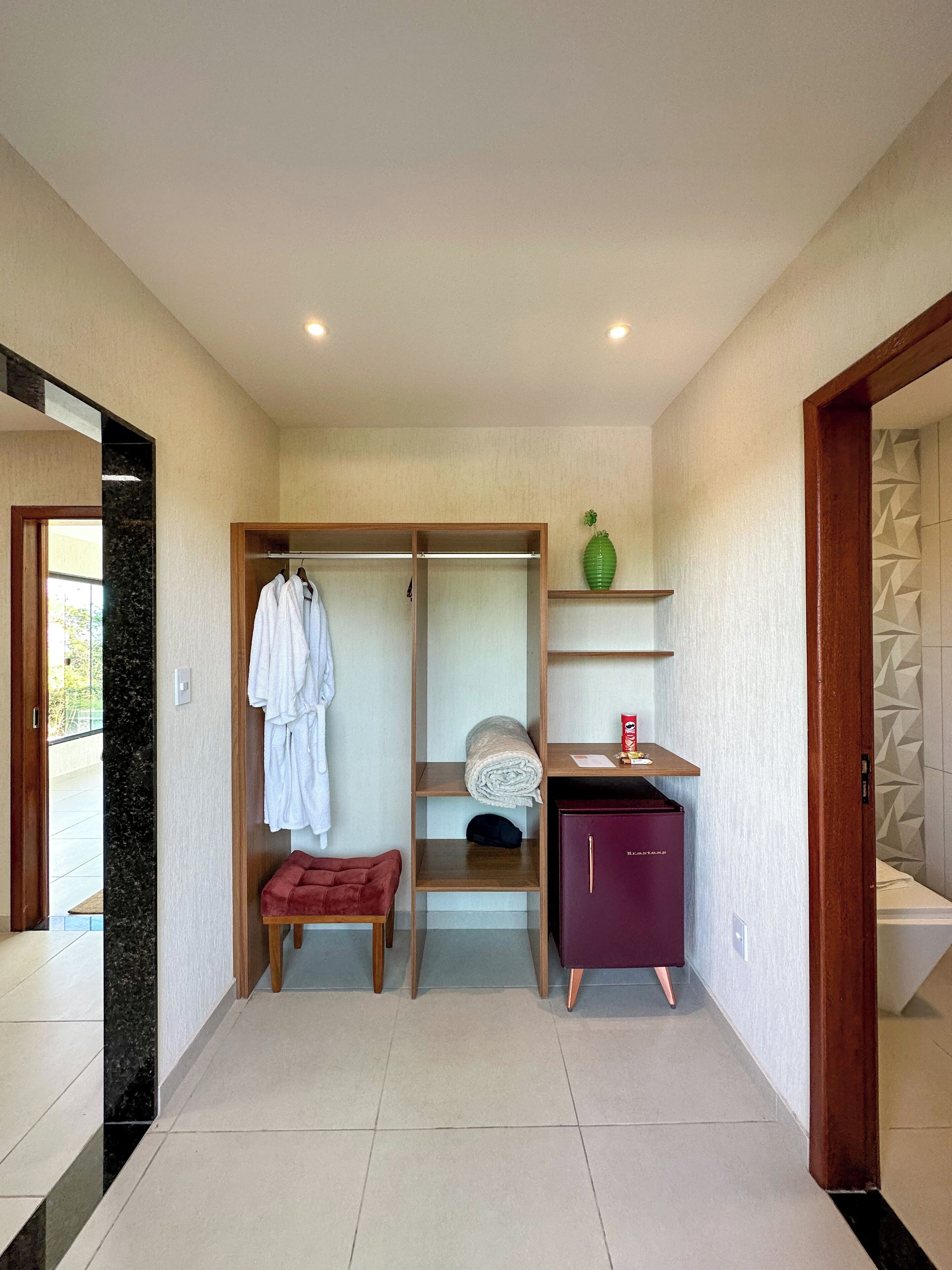 A hotel room with a walk-in closet, a red bench, a wooden shelf with a green vase, and a purple cabinet.