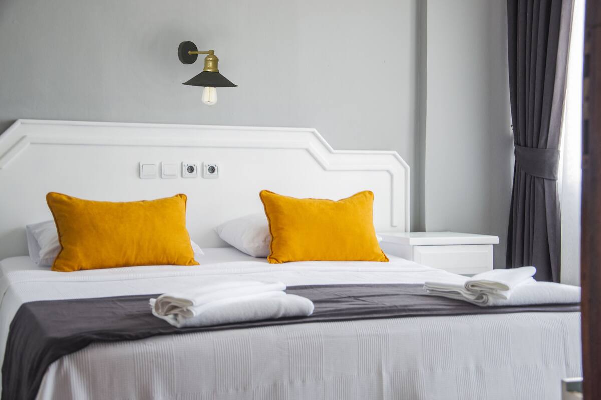 Cama bem arrumada com roupa de cama branca e duas almofadas amarelas vibrantes, uma mesa de cabeceira com abajur e uma janela com cortinas.