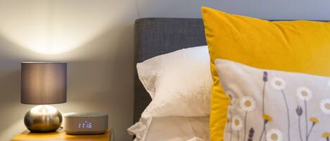 A neatly made bed with white and yellow pillows, a bedside lamp, and a digital clock on a wooden bedside table.