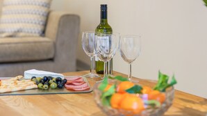 Et spisebord med et ostebord, udskårne pålæg, oliven og en skål med frugt. Der er tre vinglas og en vinflaske på bordet.