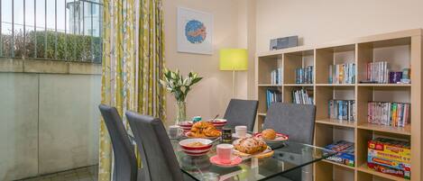 A modern dining area with a glass table, chairs, and a bookshelf filled with board games.