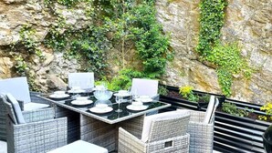 Uma área de refeições ao ar livre com móveis de vime e uma mesa posta para dois, com vista para um penhasco rochoso e vegetação.