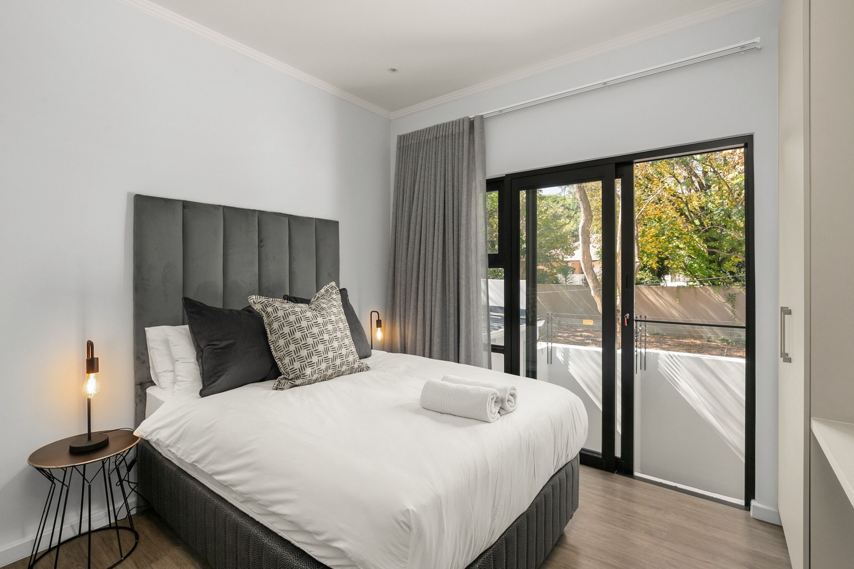 Une chambre moderne avec un grand lit, une table de chevet avec une lampe et une vue sur l’extérieur grâce à une porte-fenêtre coulissante.