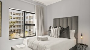 A modern bedroom with a large bed, a view of a building through a window, and a bedside table with a lamp.