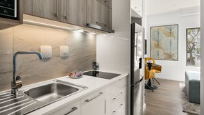 A modern kitchen with a double sink, stainless steel faucet, and a built-in oven. There is a dining area with a yellow chair and a table, and artwork on the wall.