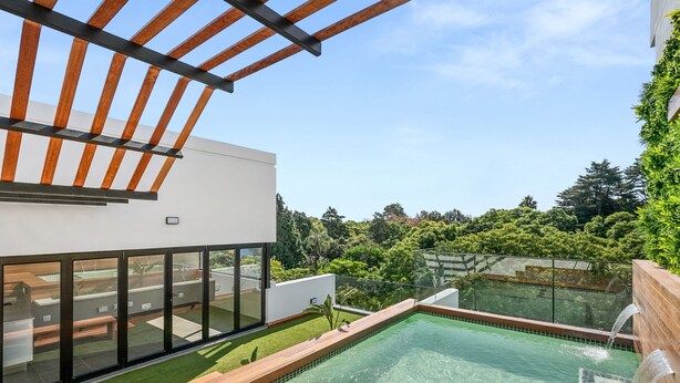 A modern outdoor pool area with a wooden deck, glass walls, and a view of a lush garden.