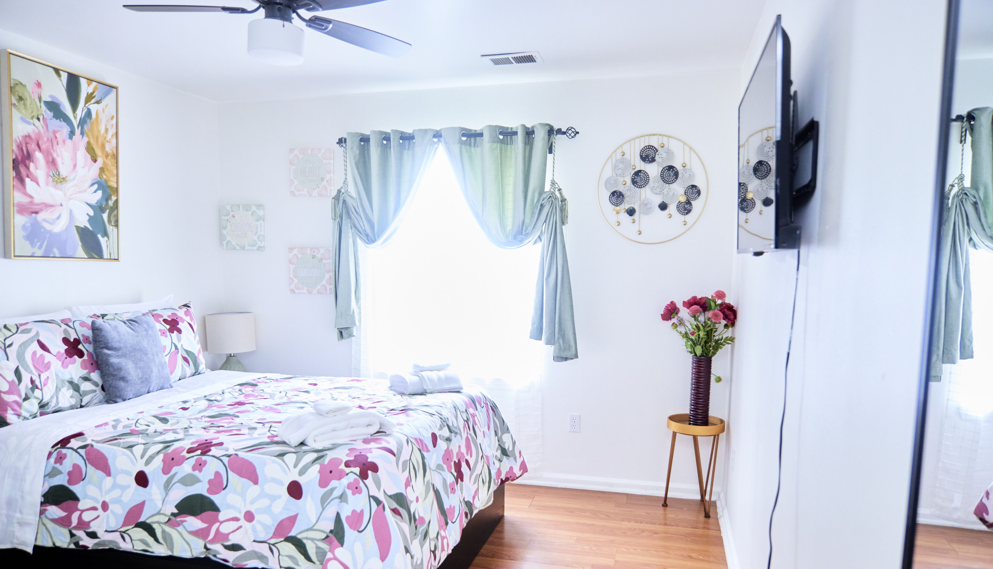 A bedroom with a bed, a ceiling fan, a window with curtains, a wall-mounted TV, and a small table with a vase of flowers.