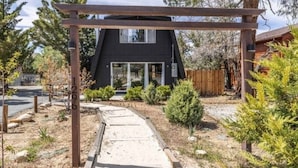 Ein modernes, schwarzes Haus mit einem Holz-Pergolen-Eingang, umgeben von einem angelegten Garten mit verschiedenen Sträuchern und einem Kiesweg.