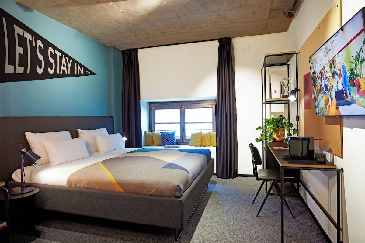 Habitación de hotel con cama, escritorio, computadora, silla, ventana con cortinas y un letrero grande en la pared.