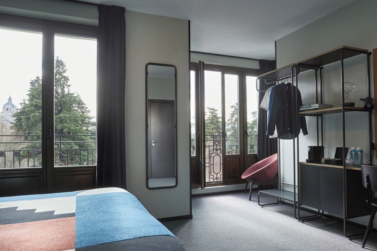 A hotel room with a bed, a desk, a chair, a wardrobe, and a large window with a view of trees and a building.