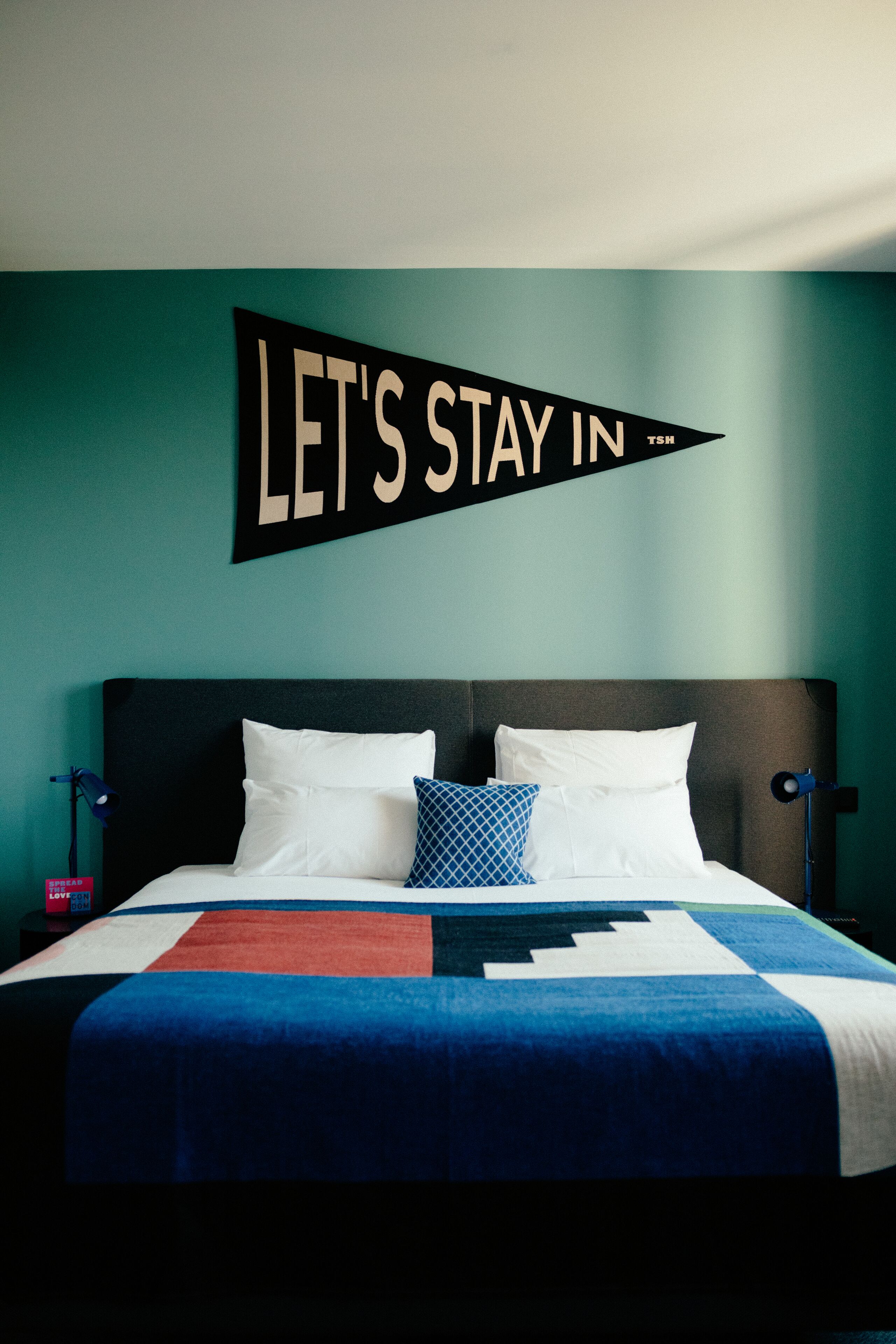 Un lit bien fait, recouvert d’une couette bleue et rouge, avec des oreillers blancs et un coussin bleu à motifs. Une pancarte au-dessus indique : « RESTONS À LA MAISON ».