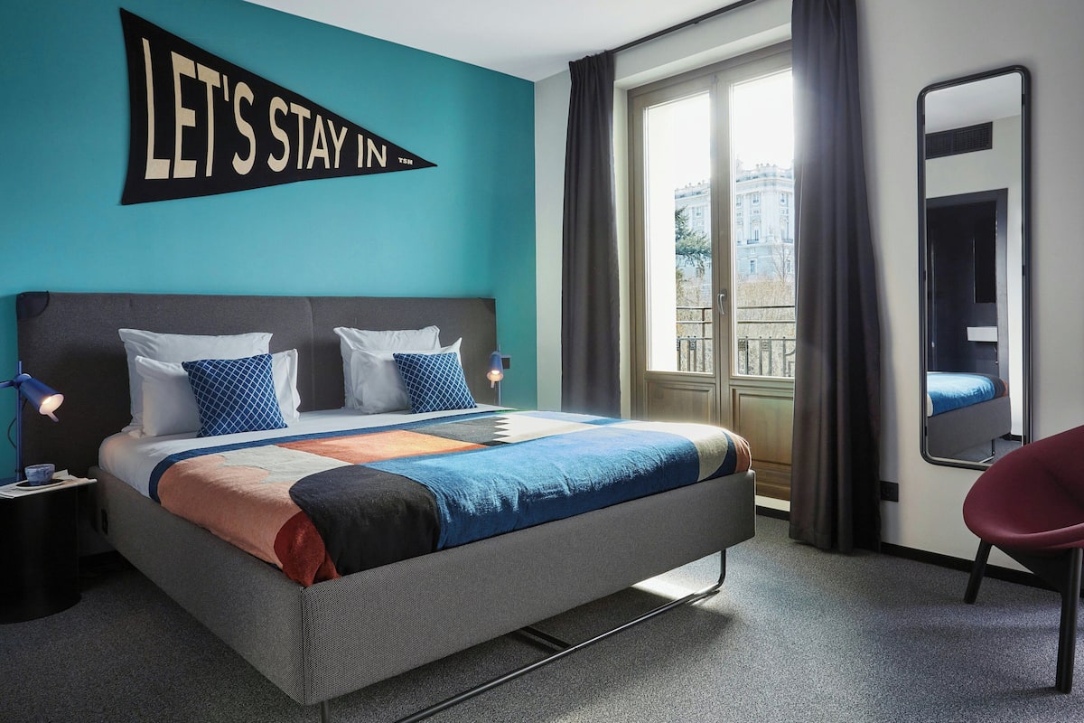 Una habitación de hotel con una cama, un cartel que dice 'LET'S STAY IN', una ventana con cortinas, un espejo y una silla.