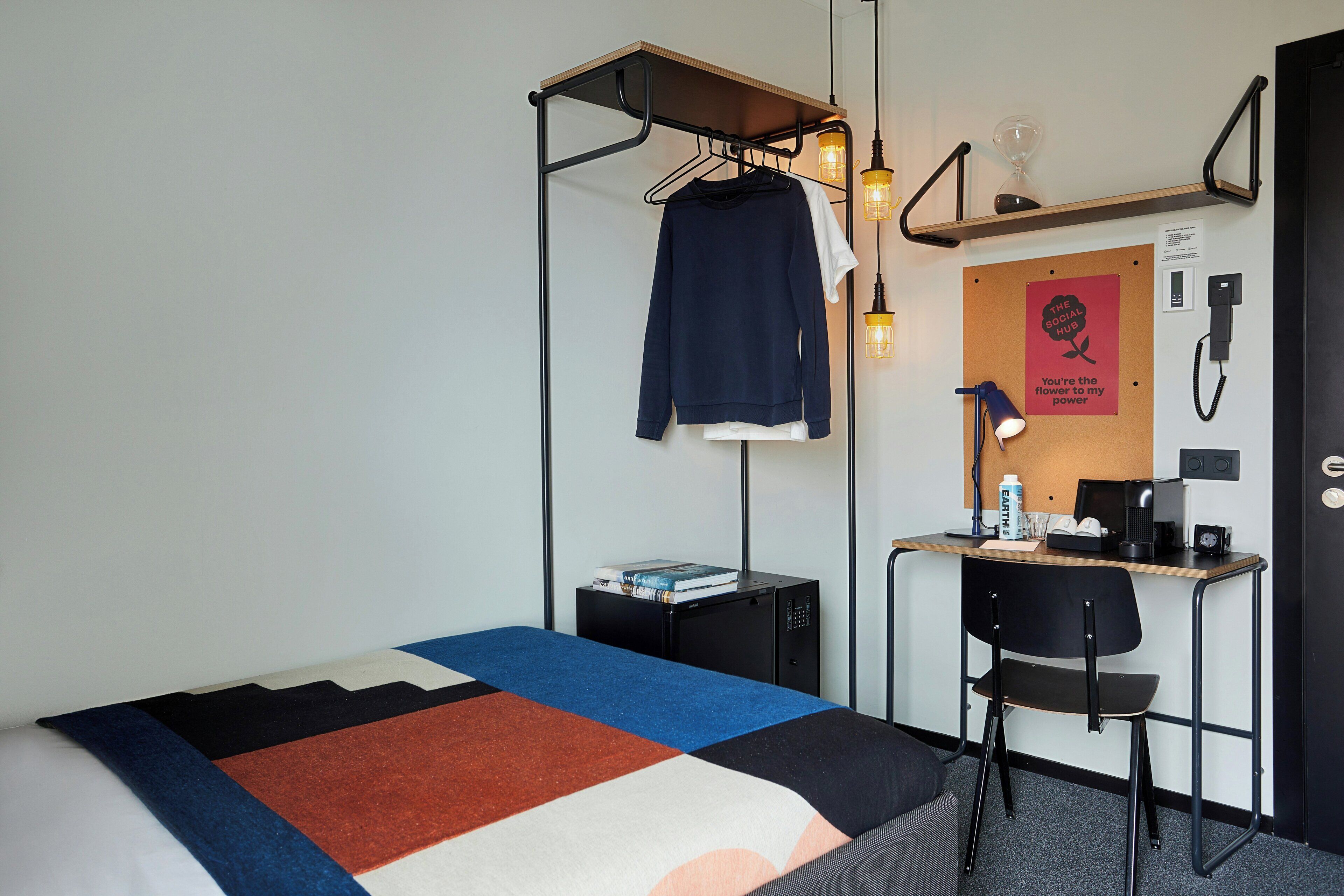 A hotel room with a bed, a desk, a chair, a hanging light fixture, and a bulletin board with a poster.