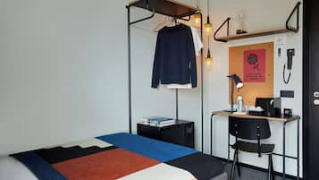 A hotel room with a bed, a desk, a chair, a hanging light fixture, and a bulletin board with a poster.