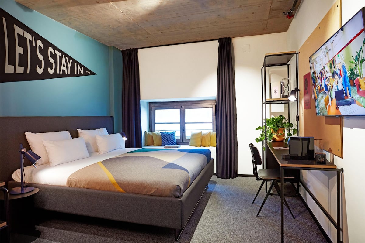 A hotel room with a bed, a desk, a chair, a large window, and a wall with a 'LET'S STAY IN' sign.