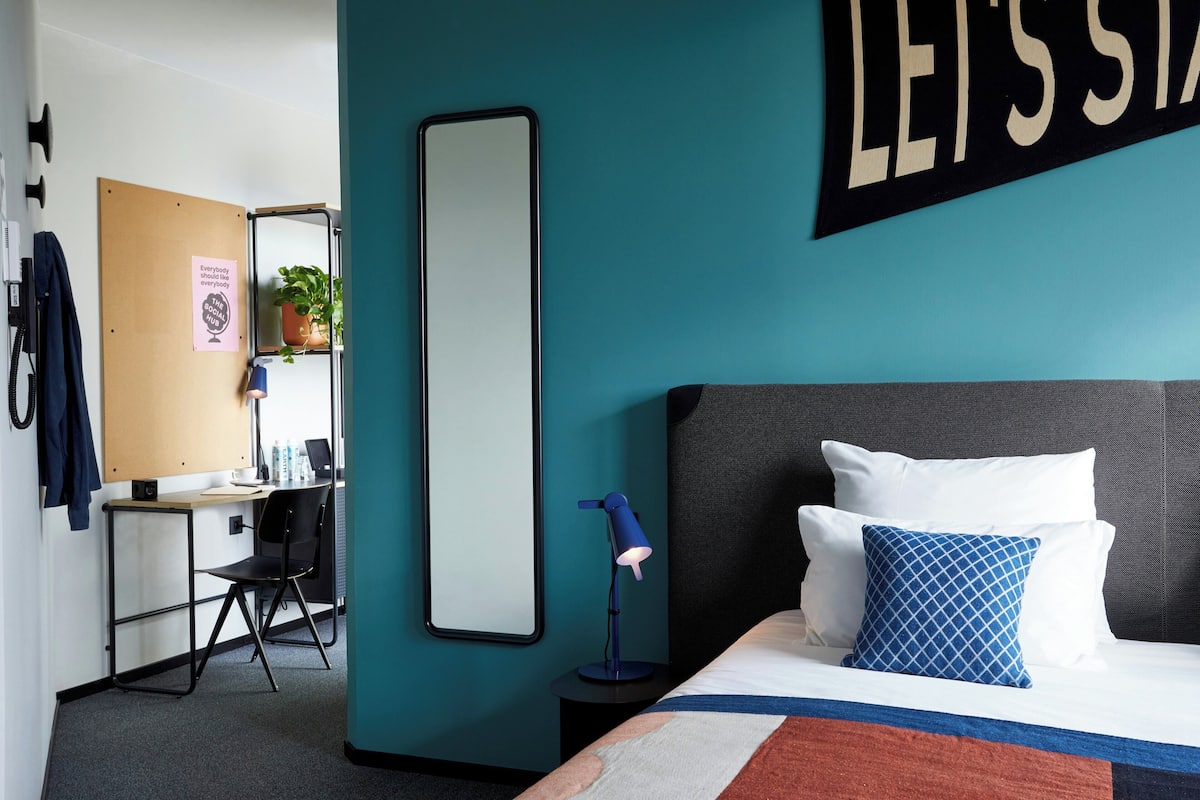 Habitación de hotel con cama, escritorio, silla, espejo y una pared con un letrero.