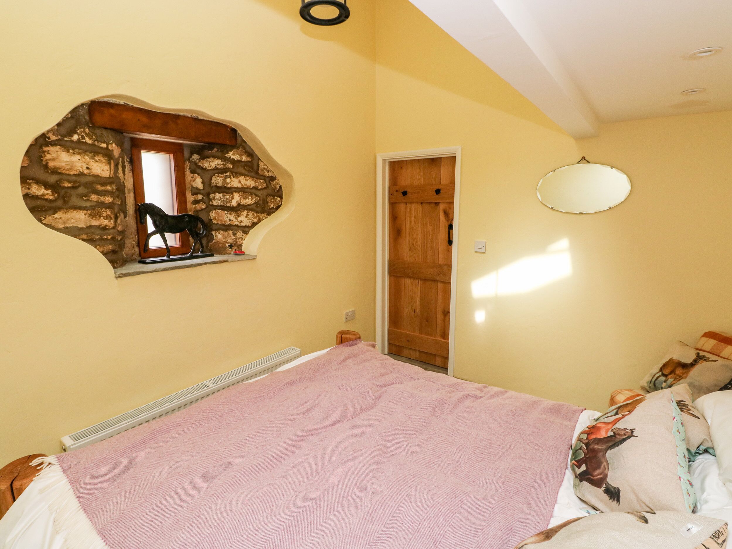 A bedroom with a bed, a wooden door, a small window with a horse decoration, and a wall-mounted mirror.