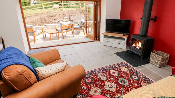 A living room with a sofa, a TV, a fireplace, and a view of an outdoor area with a fence and a tree. - Lime Kiln Cottage (Bridgend)