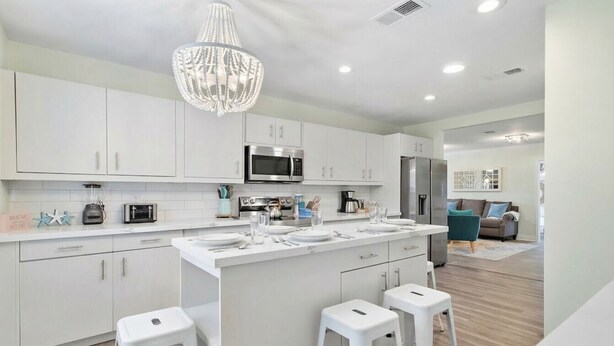 Uma cozinha moderna com armários brancos, uma ilha central com banquetas, um lustre e vista para uma sala de estar com um sofá e uma mesa de centro.