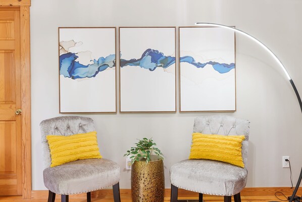 A room with two grey upholstered chairs with yellow cushions, a tall floor lamp, and three framed abstract paintings on the wall.