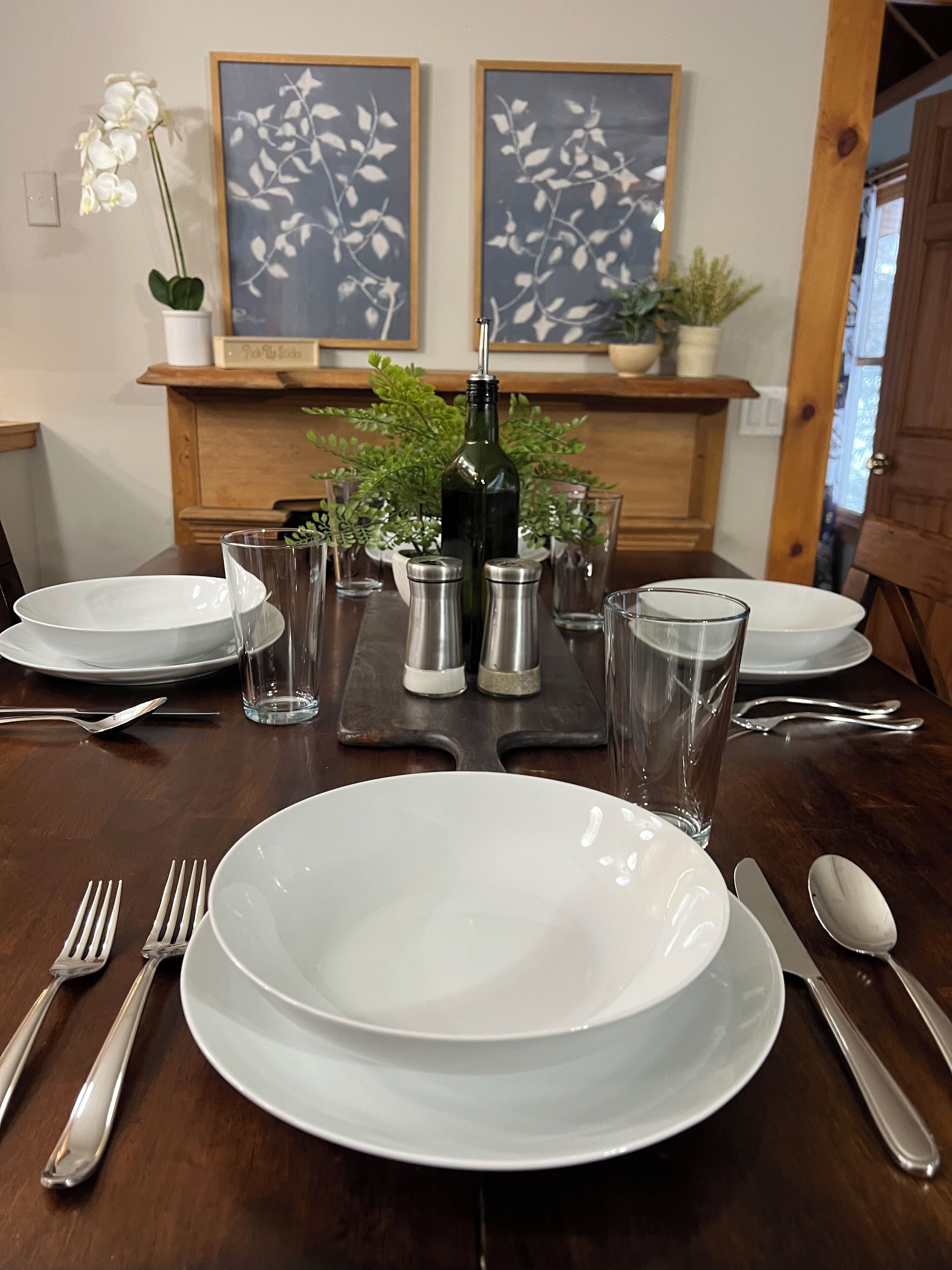 Un tavolo da pranzo apparecchiato con piatti bianchi, posate e bicchieri trasparenti, affiancato da due quadri floreali incorniciati e una pianta in vaso.