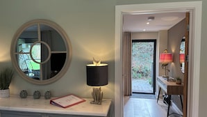 A room with a round mirror, a lamp, a small table with decorative items, and a view of a garden through an open door. - Primrose (Bodmin)