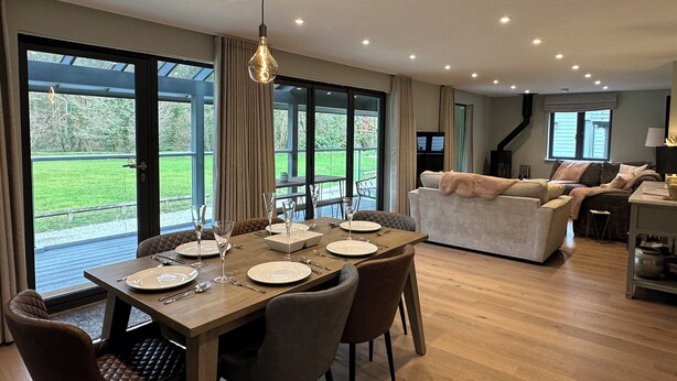 A spacious dining area with a wooden table set for six, large windows offering a view of the outdoors, and a cozy living area with a sofa and a television. - Primrose (Bodmin)