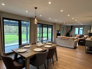 A spacious dining area with a wooden table set for six, large windows offering a view of the outdoors, and a cozy living area with a sofa and a television. - Primrose (Bodmin)