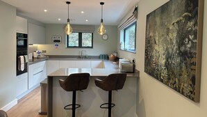A modern kitchen with a central island, two bar stools, pendant lighting, a large window, and a painting on the wall. - Primrose (Bodmin)