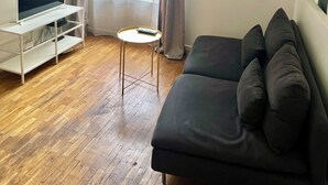 A living room with a TV on a white stand, a small round table with a remote, a dark sofa, wooden flooring, and a window with curtains.