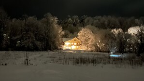 A snow-covered landscape with a house illuminated by warm lights, surrounded by trees. - Village Home with 15 acre backyard on the river (Cavendish)