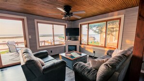 A modern living room with leather sofas, a coffee table, and a TV. Large windows offer views of the beach and boats.