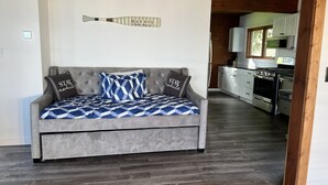 A grey sofa with patterned cushions, a wall-mounted baseball bat, and a kitchen in the background.