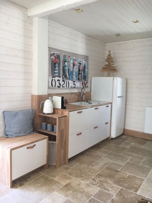 Une cuisine compacte avec un réfrigérateur blanc, un évier et des placards en bois. Un tableau représentant des phares est accroché au mur, et un petit tabouret en bois est posé à proximité.
