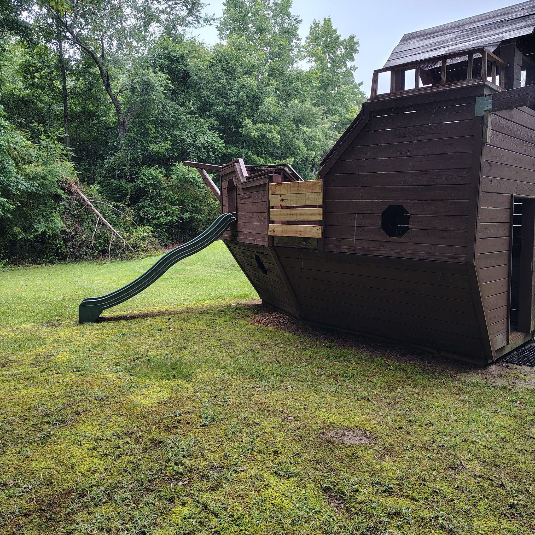 A wooden play structure with a slide and climbing area, surrounded by grass and trees.