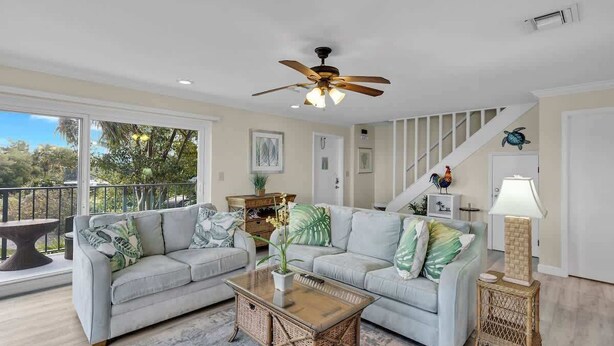 A spacious living room with two light grey sofas, a wooden coffee table, and a ceiling fan. A staircase with white railings leads to another level.