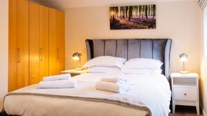 A neatly made bed with white linens and towels, a grey upholstered headboard, and a painting of a forest scene above it.