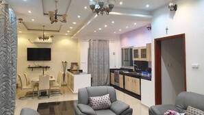 A modern living room with a grey sofa, a dining area with a white table and chairs, a kitchen with wooden cabinets, and a TV mounted on the wall. - Magnificent house in quiet residential area (Yaoundé)