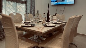 A dining area with a marble table set for six, surrounded by beige upholstered chairs. A television mounted on the wall displays a scenic landscape. - Magnificent house in quiet residential area (Yaoundé)