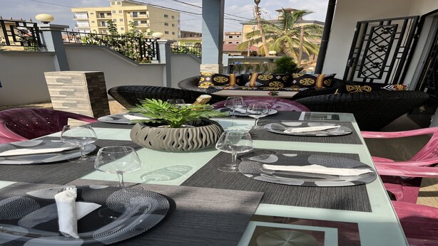 An outdoor dining area with a table set for four, surrounded by wicker chairs, a decorative plant, and a view of the neighborhood. - Magnificent house in quiet residential area (Yaoundé)