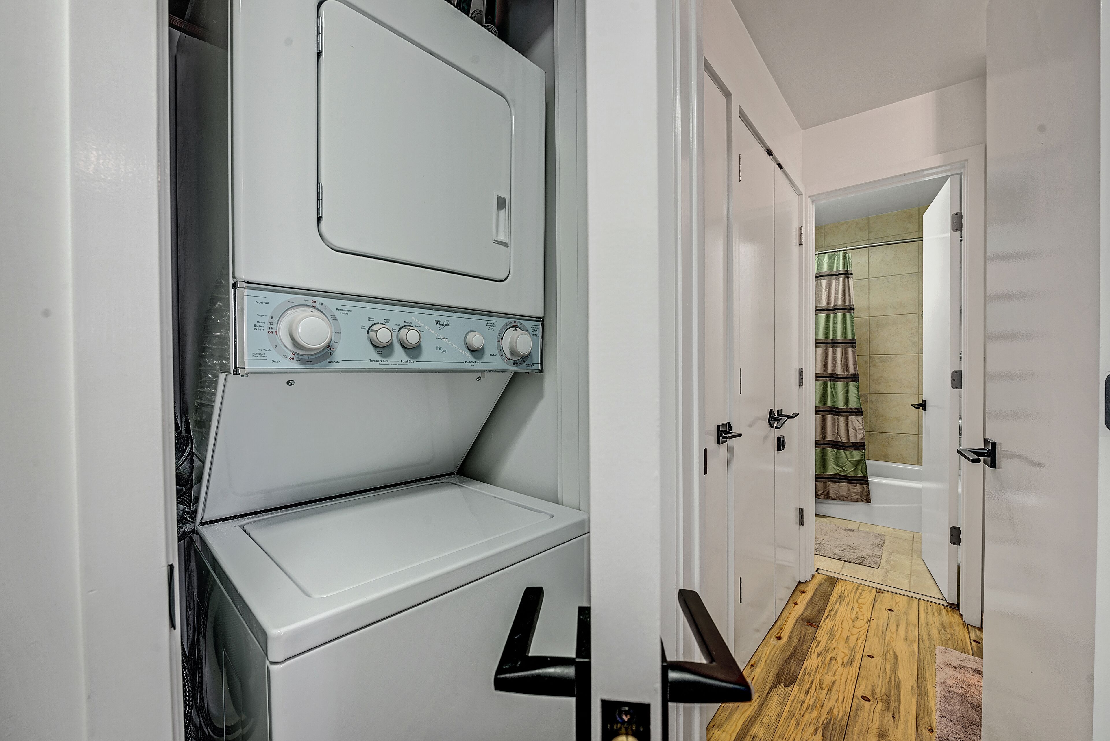 A laundry room with stacked washers and dryers, a wooden floor, and a door leading to a bathroom with a shower and towel rack.