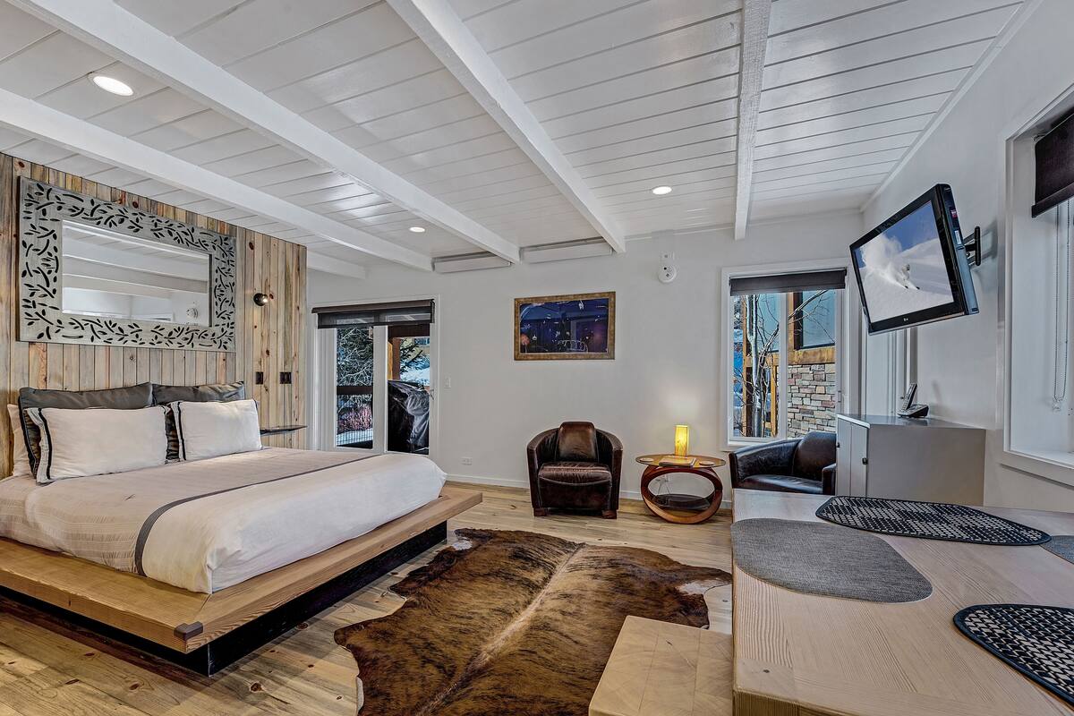 A modern bedroom with a large bed, a wooden headboard, a mounted TV, and a view of the outdoors.