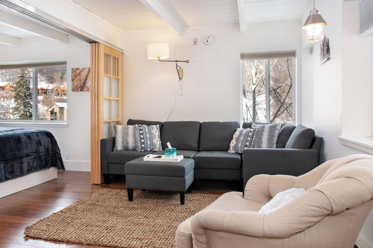A modern living room with a sectional sofa, a beige armchair, a small ottoman, and a large window offering a view of a snowy landscape.