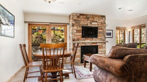 A living room with a stone fireplace, a TV mounted above it, a brown sofa, and a dining area with wooden chairs and a table. - Willows Condos by Snowmass Vacations (Snowmass Village)