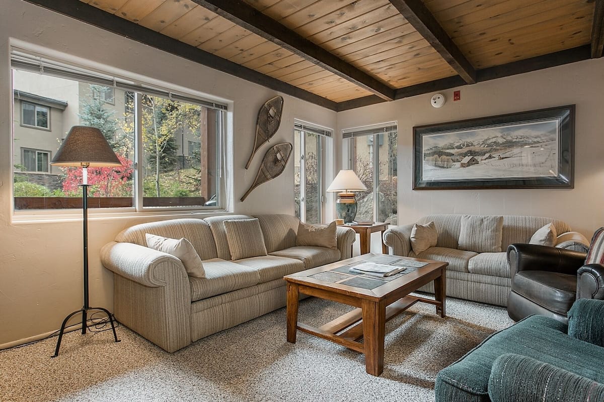 A cozy living room with a wooden ceiling, large windows, a sofa, a coffee table, and a painting on the wall.