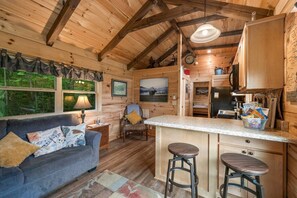 Interior de una cabaña con sofá, sillón, zona de cocina y mesa de comedor.