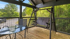 Un espacio al aire libre techado con un piso de madera, una mesa de vidrio, una hamaca y vistas a árboles.