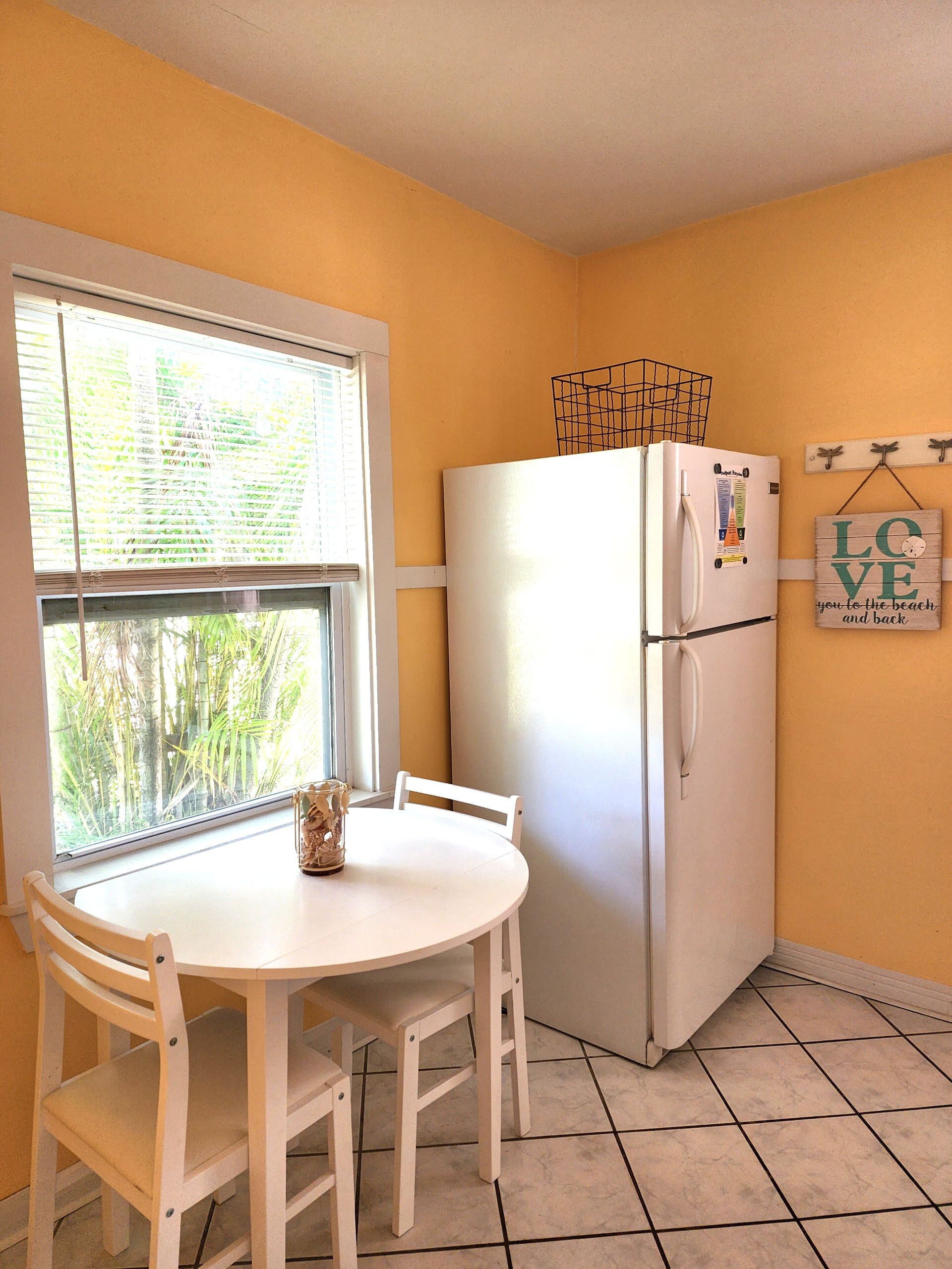 Una cocina con un refrigerador blanco, una ventana con persianas, una mesa blanca con sillas y una pared con un letrero que dice 'LOVE'.